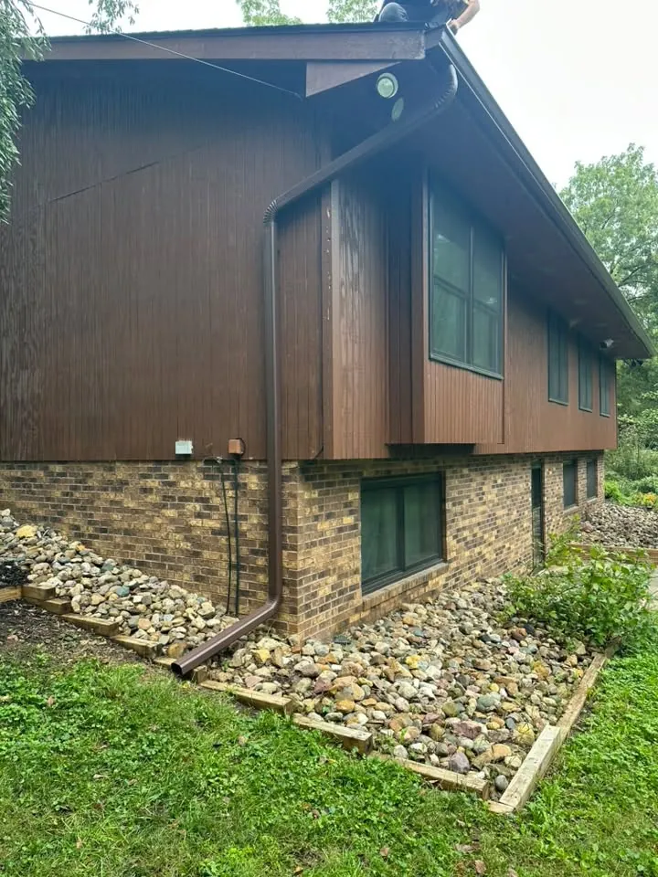 Brown gutters and downspout on a split-level home with brown wood siding and a rock bed
