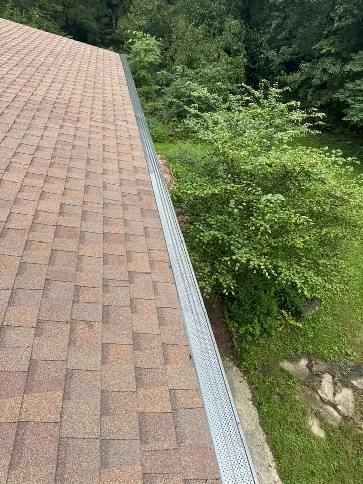Long white gutter run along a shingled roof edge above a wooded slope